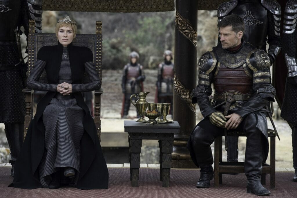 A blonde woman is sitting on a throne, a knight is sitting next to her; some guards stand behind them.