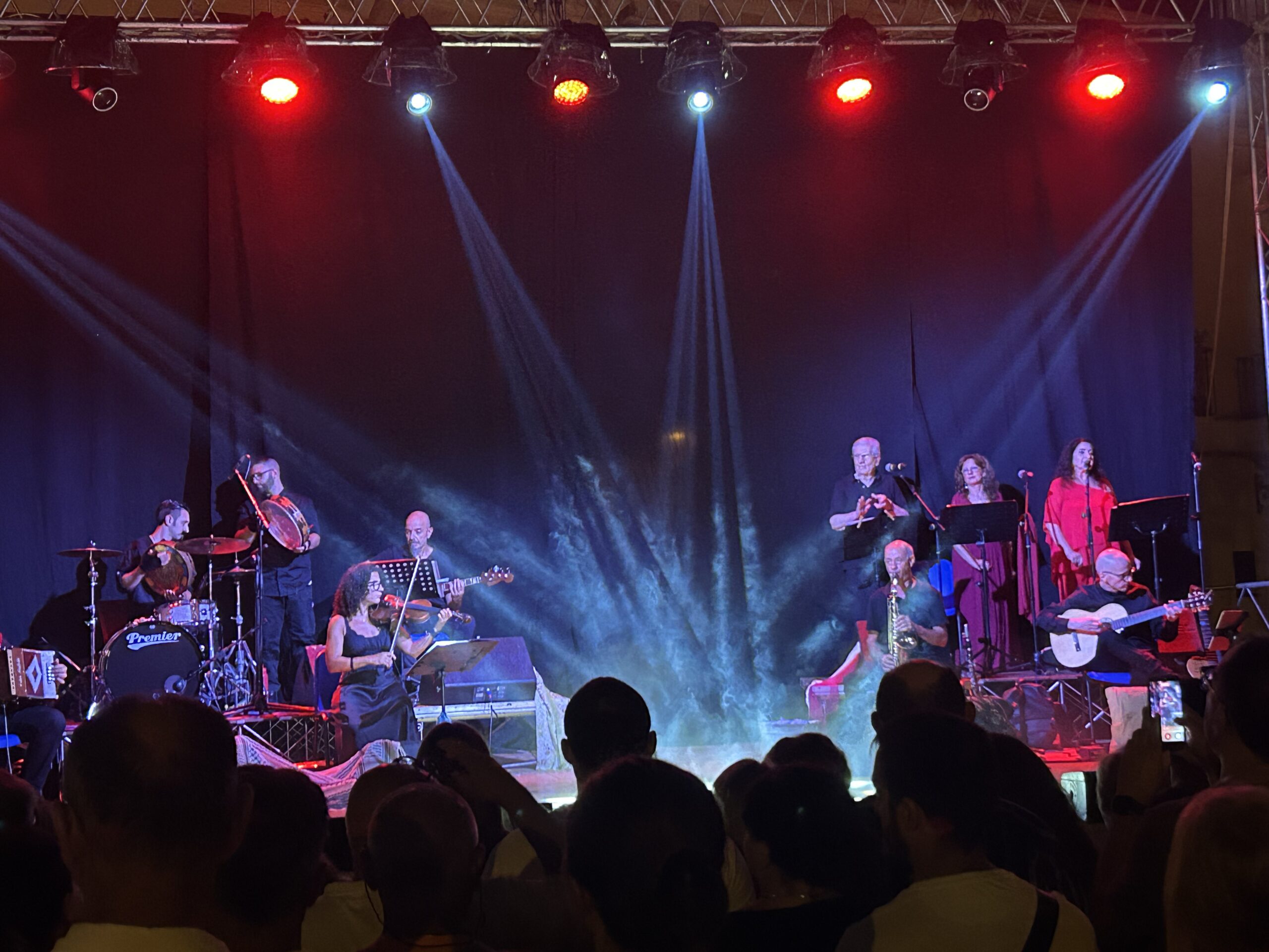 Band Taricata playing pizzica on stage. On the left, there are drummers, a violinist, a semi-acoustic guitar player. On the right, there are 3 singers, a saxophone player and the second acoustic guitar player.