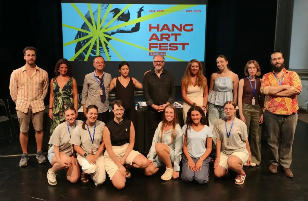 The Hangartfest 2026 team posing on stage during the press conference in Pesaro, including artistic director Antonio Cioffi and board members Paolo Paggi and Masako Matsushita, in front of the festival's graphic screen.